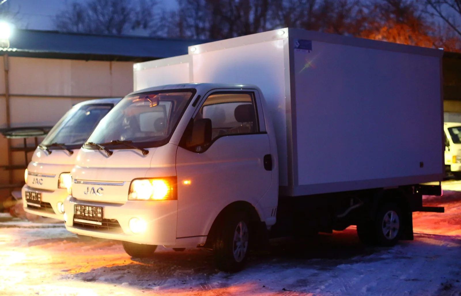 JAC X200 шассиіндегі изотермиялық фургон — партияның екі көлігі алдынан, шамдары қосулы — COND